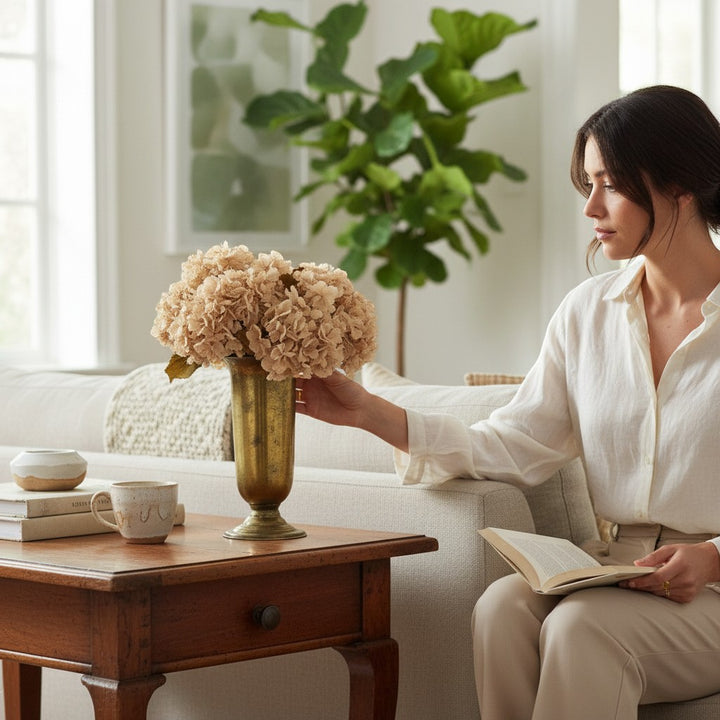 Chic Collective 24" beige hydrangea spray in a brass vase on a wooden side table, styled with books and a mug.
