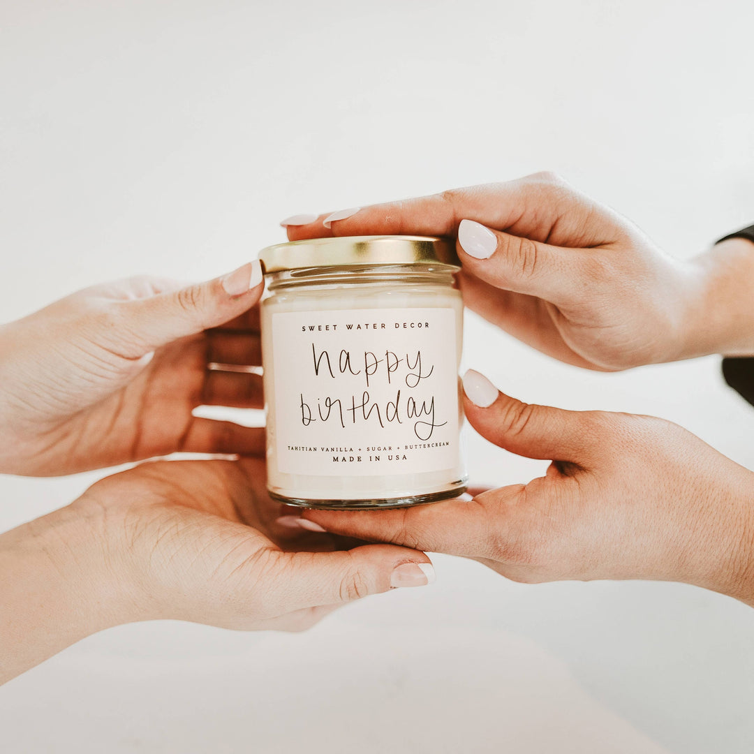 Chic Collective Happy Birthday vanilla buttercream scented soy candle in a clear jar with gold lid, held by two people.