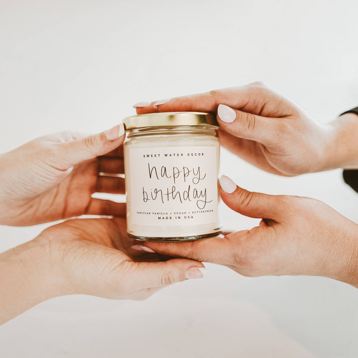Chic Collective Happy Birthday vanilla buttercream scented soy candle in a clear jar with gold lid, held by two people.