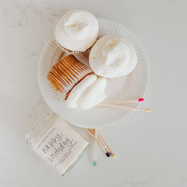 Chic Collective Happy Birthday vanilla buttercream scented soy candle in a clear jar with a white label. Three frosted cupcakes on a white plate and colorful matches are nearby.