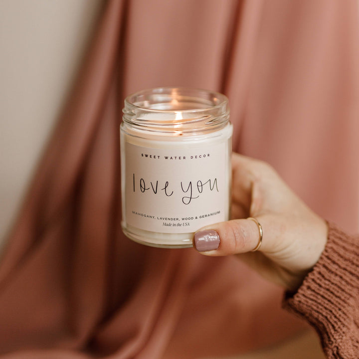 Chic Collective Love You 9 oz soy candle with driftwood and mahogany scent, held by a hand with a gold ring.