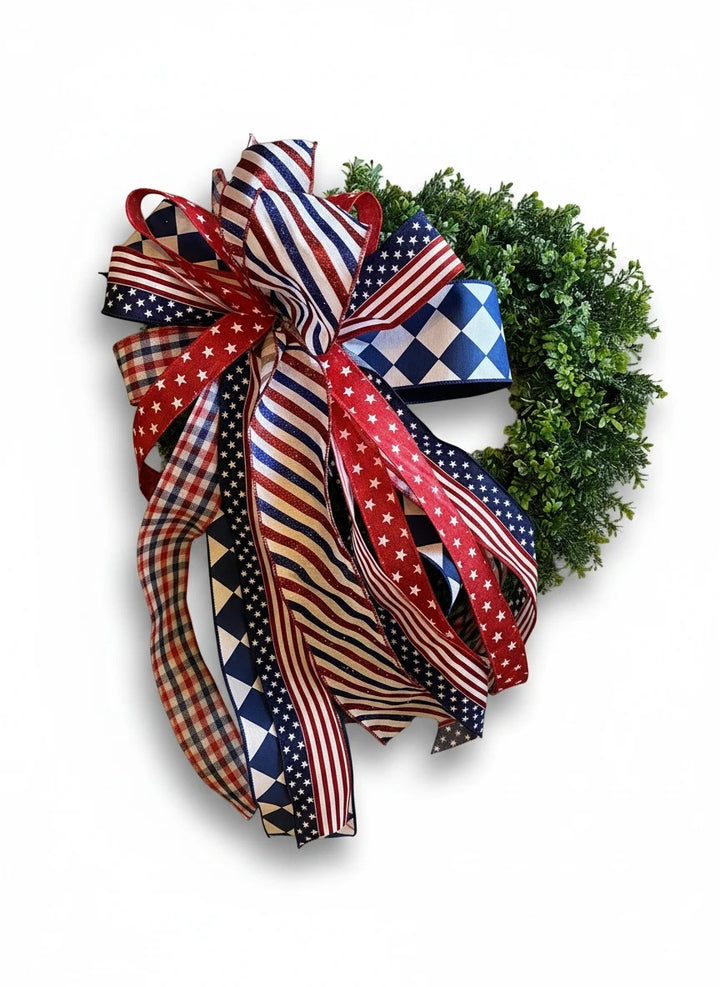 Chic Collective patriotic wreath with a lush green base and a large bow made of red, white, and blue ribbons featuring stars, stripes, and checkered patterns.