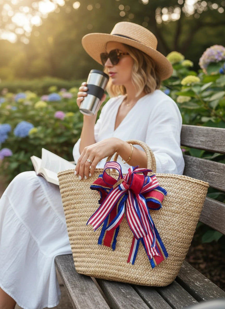 Chic Collective straw tote bag with a red, white, and blue striped bow, sitting on a park bench with a woman.