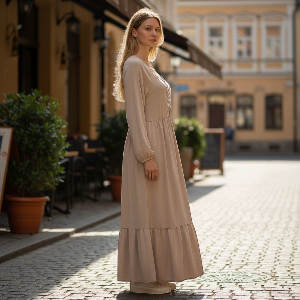 Chic Collective beige ruched maxi dress with long sleeves and tiered hem, worn by a model on a cobblestone street.