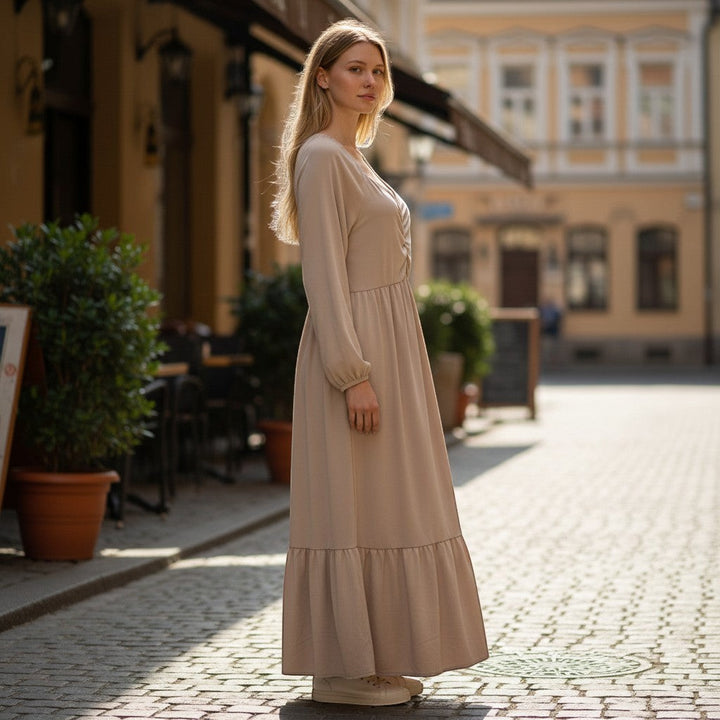 Chic Collective beige ruched maxi dress with long sleeves and tiered hem, worn by a model on a cobblestone street.