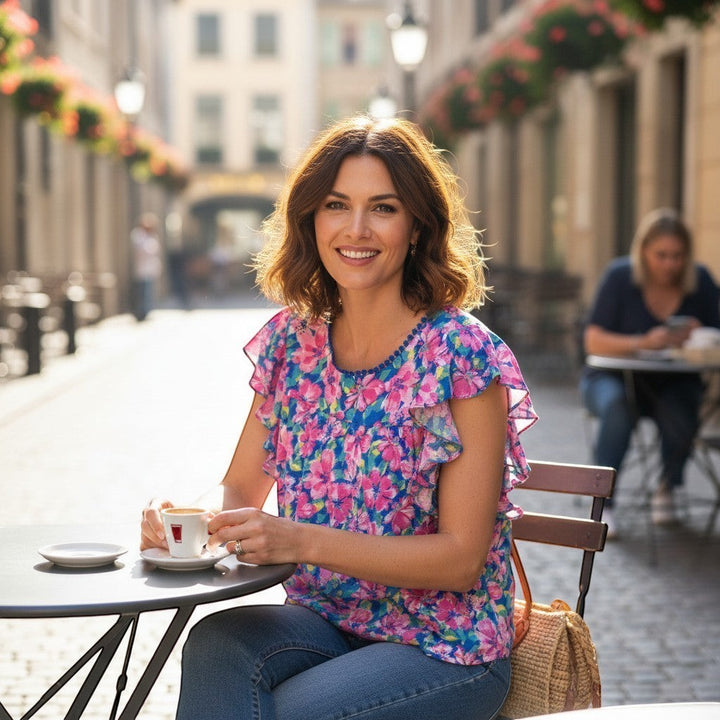 Chic Collective blue and pink hibiscus floral ruffle top with short sleeves and a round neckline, worn by a woman sitting at a cafe.