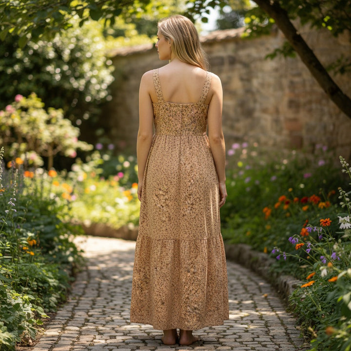 Chic Collective floral tan maxi dress with tiered skirt and square neckline, worn by a woman standing on a cobblestone path in a garden.