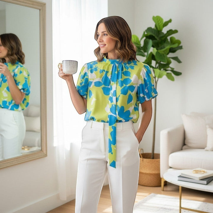 Chic Collective lime green and blue floral print blouse with short sleeves and a tie neck detail, worn by a woman in white pants.
